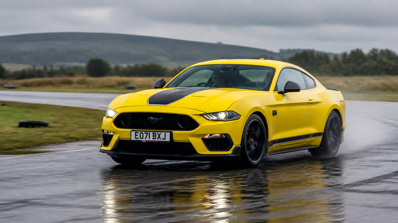 Carro esportivo amarelo Ford Mustang em pista molhada, com céu nublado e relevo ao fundo.