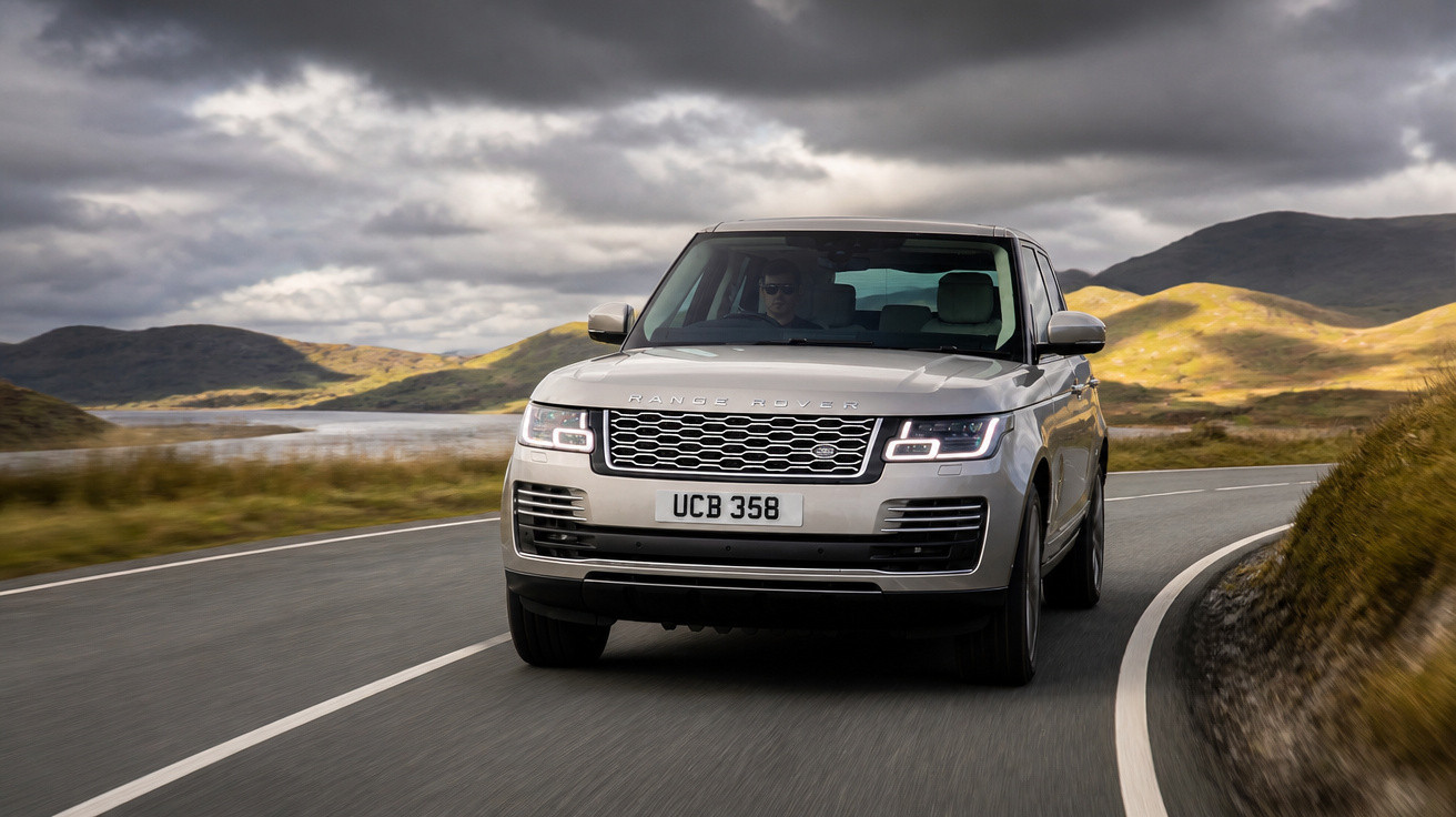 Carro SUV Range Rover prata dirigindo em estrada sinuosa com colinas e céu nublado ao fundo.